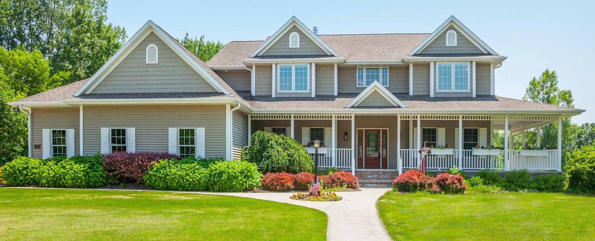 A large two-story home with pale siding, a wraparound porch and a clean walkway on a bright summer day
